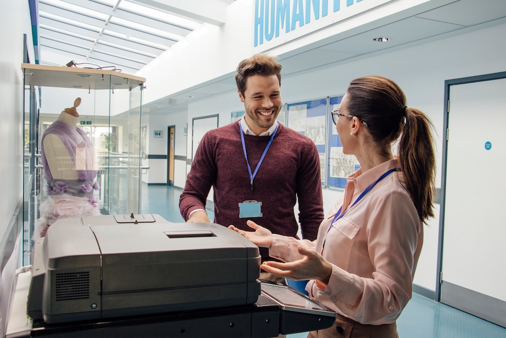 Two teachers are talking while standing at a printer in the school hall.