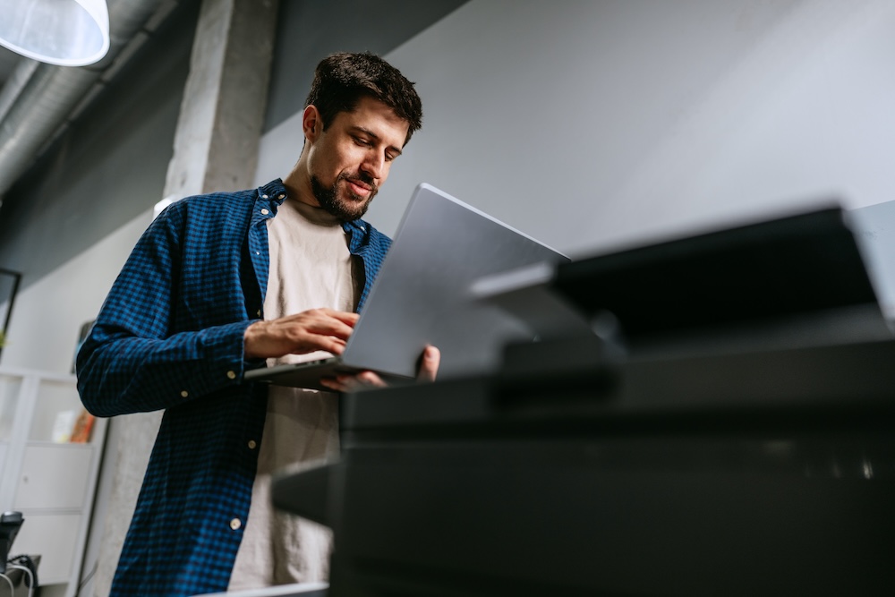 A man stands in a contemporary office environment, focused on his laptop while typing. The space features minimalistic decor and a printer nearby, fostering productivity.