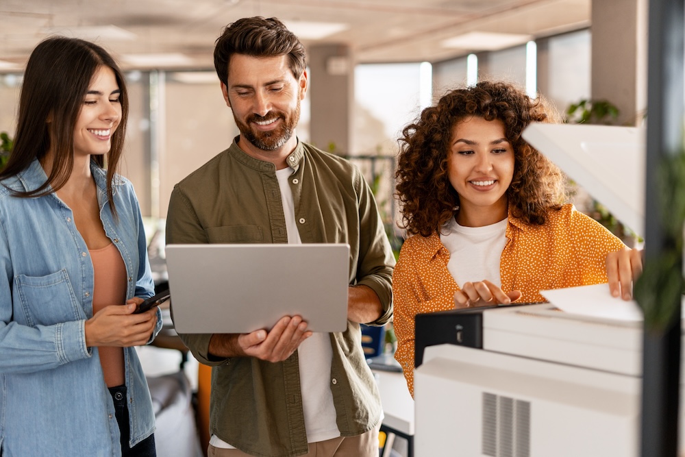 Small business employees reviewing office printing activity and documents beside a multifunction office printer, highlighting managed print services, print cost management, and modern workplace technology.